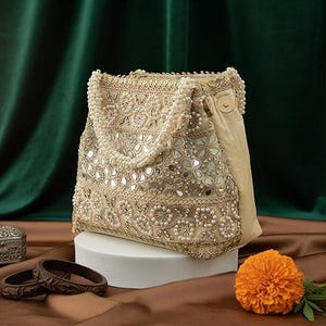 K&ouml;nnte beinhalten: Eine beige Handtasche mit aufwendiger Stickerei, Spiegelakzenten und Perlenverzierungen. Die Tasche hat einen perlenbesetzten Griff und einen satinartigen Stoff an der Seite. Eine orangefarbene Blume und braune Armreifen sind ebenfalls zu sehen.