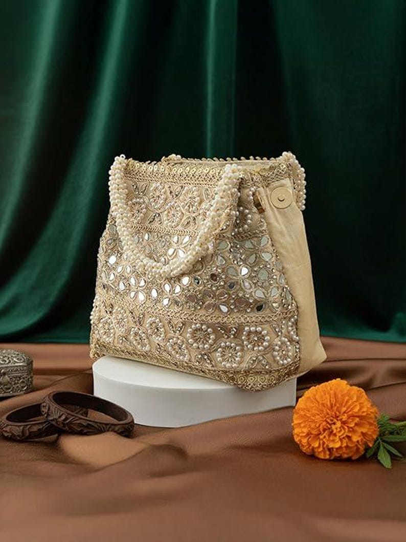 K&ouml;nnte beinhalten: Eine beige Handtasche mit aufwendiger Stickerei, Spiegelakzenten und Perlenverzierungen. Die Tasche hat einen perlenbesetzten Griff und einen satinartigen Stoff an der Seite. Eine orangefarbene Blume und braune Armreifen sind ebenfalls zu sehen.