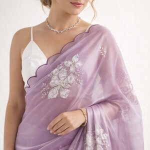 May include: A sheer, lavender saree with intricate silver floral embroidery. The saree features a scalloped edge with a dark purple trim. A white camisole is visible underneath. A delicate diamond necklace and bracelet complete the look.