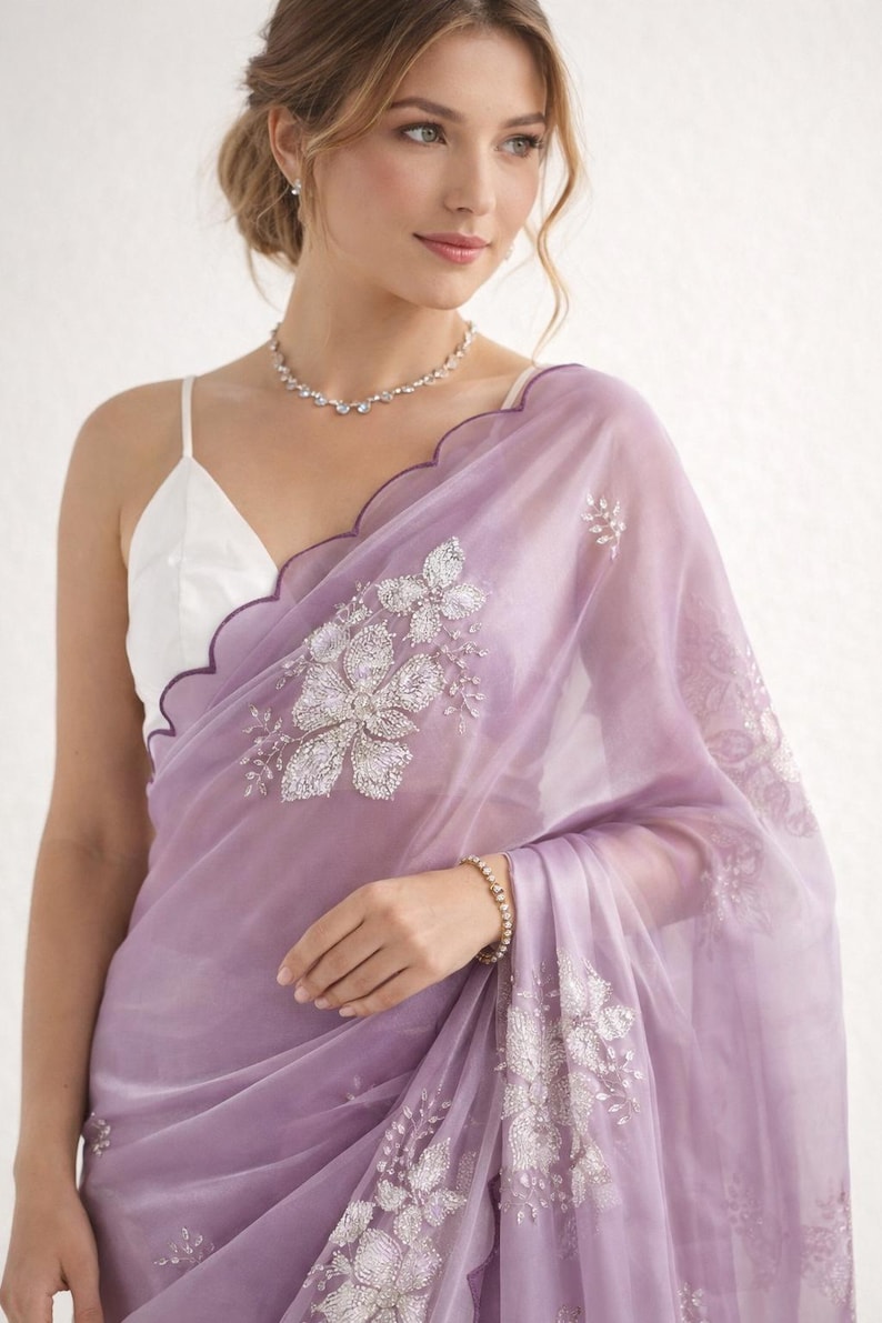May include: A sheer, lavender saree with intricate silver floral embroidery. The saree features a scalloped edge with a dark purple trim. A white camisole is visible underneath. A delicate diamond necklace and bracelet complete the look.