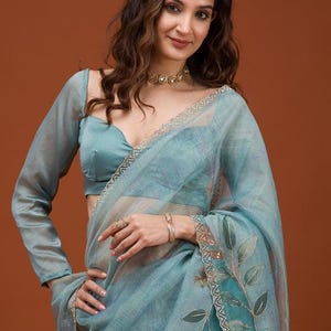 May include: A light blue sheer saree featuring a delicate hand-painted leaf pattern in shades of green and brown along the border, accented with a metallic silver trim. The ensemble includes a matching long-sleeved blouse with a sweetheart neckline.