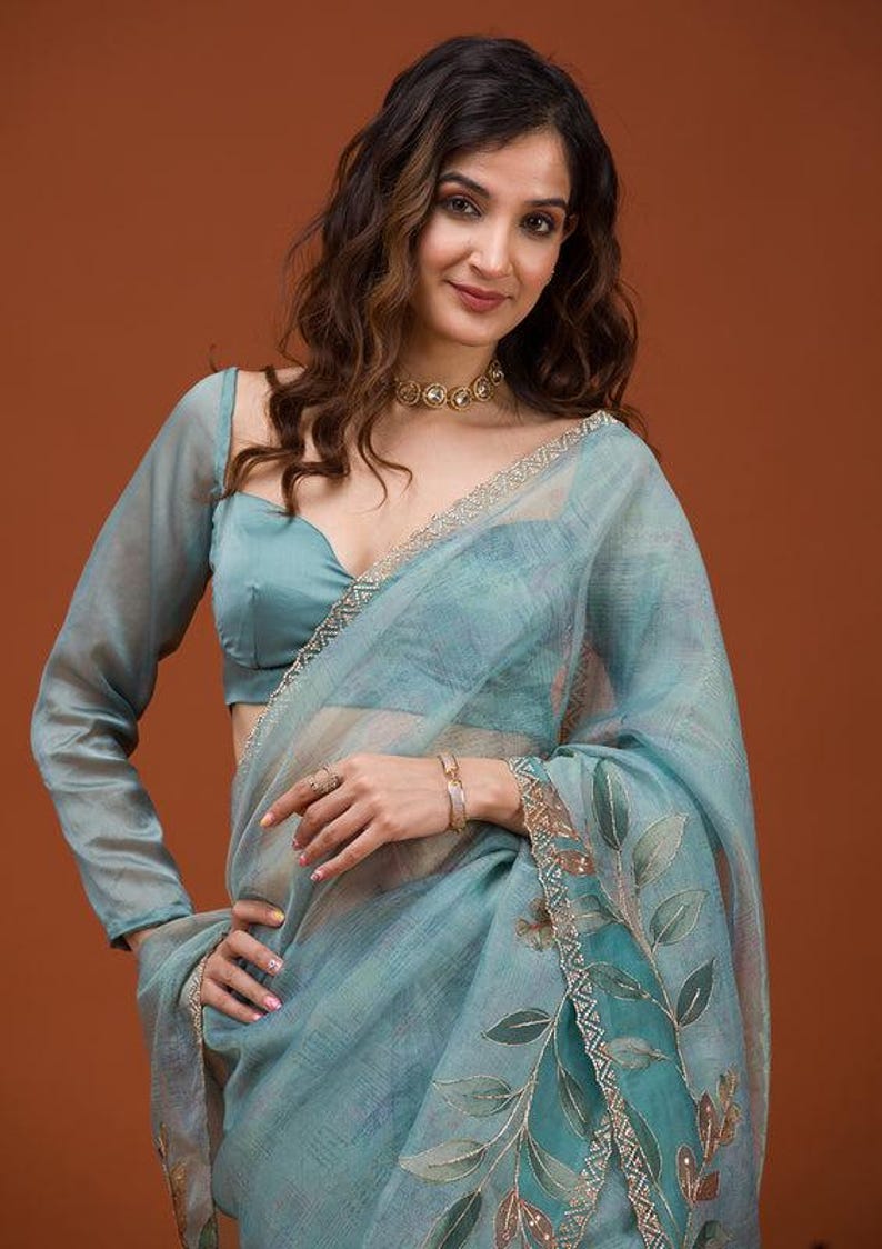 May include: A light blue sheer saree featuring a delicate hand-painted leaf pattern in shades of green and brown along the border, accented with a metallic silver trim. The ensemble includes a matching long-sleeved blouse with a sweetheart neckline.