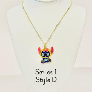 K&ouml;nnte beinhalten: Goldkette mit einem Anh&auml;nger in Form von Stitch, einem blauen und gelben Charakter. Der Anh&auml;nger hat rosa Akzente und eine blaue Schleife. Die Kette ist mit "Serie 1 Style D" beschriftet.