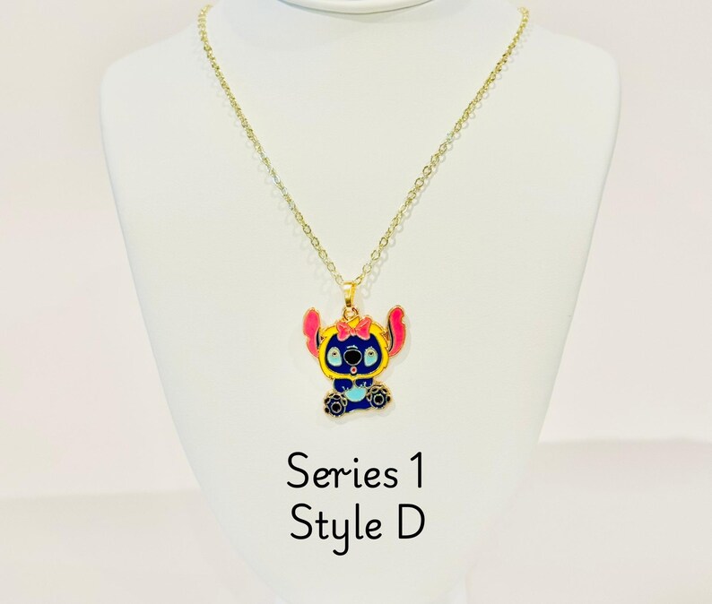 K&ouml;nnte beinhalten: Goldkette mit einem Anh&auml;nger in Form von Stitch, einem blauen und gelben Charakter. Der Anh&auml;nger hat rosa Akzente und eine blaue Schleife. Die Kette ist mit "Serie 1 Style D" beschriftet.
