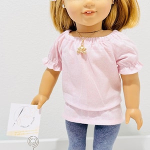 May include: A doll with blonde hair and blue eyes, wearing a pink top, gray leggings, and white sneakers. The doll has a gold necklace and bows in her hair. A small sign is held in the doll's hand, and a pink rose is in a small pot.