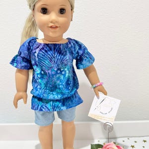 May include: A doll wearing a blue patterned top with ruffled sleeves and denim shorts. The doll has blonde hair and brown sandals. A tag that says "Stellar Styles" is held in the doll's hand.