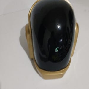 Może przedstawiać: Czarny i złoty kask z błyszczącym wykończeniem. Kask ma zaokrągloną g&oacute;rę oraz złote wykończenia i akcenty. Projekt przypomina futurystyczny lub elektroniczny motyw muzyczny.