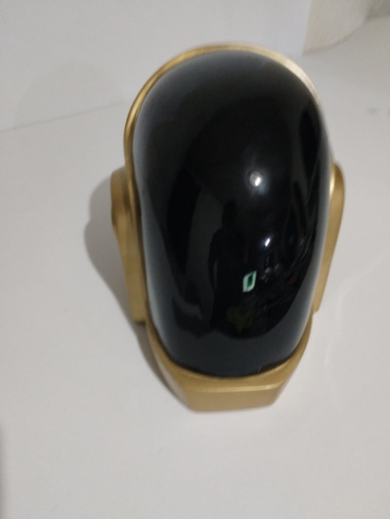 Może przedstawiać: Czarny i złoty kask z błyszczącym wykończeniem. Kask ma zaokrągloną g&oacute;rę oraz złote wykończenia i akcenty. Projekt przypomina futurystyczny lub elektroniczny motyw muzyczny.