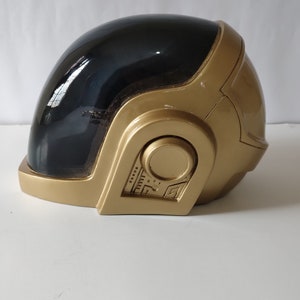 May include: A gold and black helmet with a visor and a circular panel with numbers and symbols. The helmet is made of plastic and has a glossy finish.