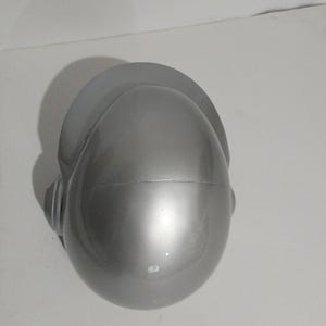 Może przedstawiać: Srebrny kask z daszkiem i nausznikami. Kask ma gładką, odblaskową powierzchnię i zaokrąglony kształt. Daszek to płaski, zakrzywiony element wystający z g&oacute;ry kasku.