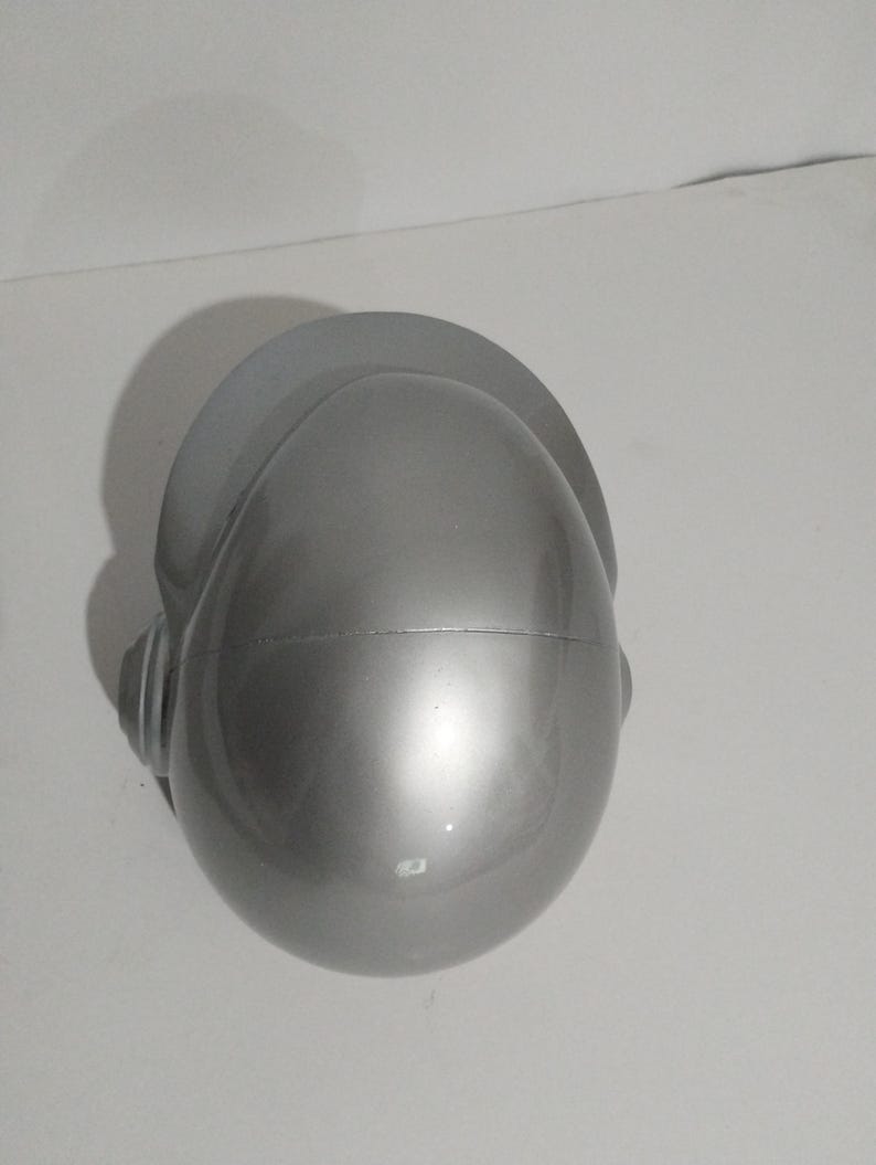 Może przedstawiać: Srebrny kask z daszkiem i nausznikami. Kask ma gładką, odblaskową powierzchnię i zaokrąglony kształt. Daszek to płaski, zakrzywiony element wystający z g&oacute;ry kasku.