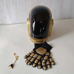 Puede incluir: Un casco negro y dorado con una cadena y un colgante dorados. El casco tiene una visera negra y detalles dorados. El colgante es un triángulo dorado. El casco está sobre un fondo blanco.