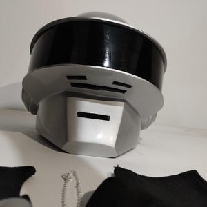 Może przedstawiać: Futurystyczny hełm w odcieniach szarości i czerni. Hełm ma zaokrągloną g&oacute;rę z czarnym wizjerem i prostokątnym otworem na usta. Projekt obejmuje poziome linie i srebrne wykończenie, sugerując motyw science fiction lub cosplay.