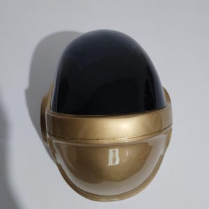 Może przedstawiać: Złoty kask z czarnym wizjerem. Kask ma zaokrąglony kształt z gładką, odblaskową powierzchnią. Projekt przypomina futurystyczny lub elektroniczny motyw muzyczny.