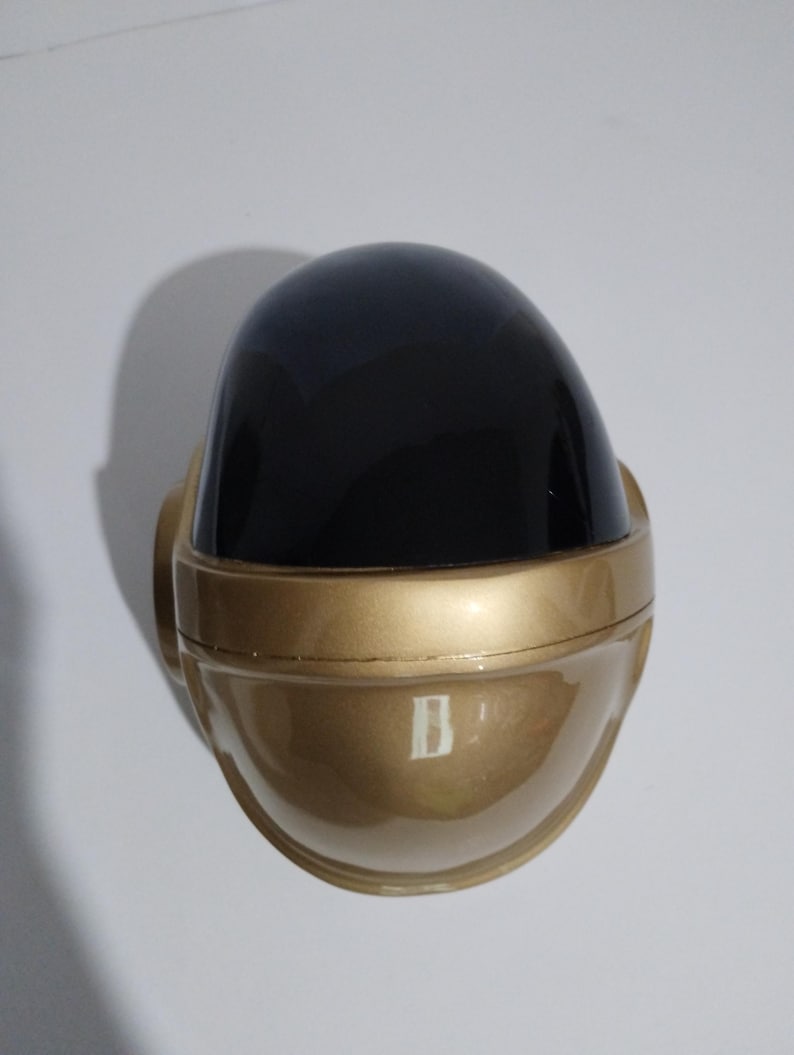 Może przedstawiać: Złoty kask z czarnym wizjerem. Kask ma zaokrąglony kształt z gładką, odblaskową powierzchnią. Projekt przypomina futurystyczny lub elektroniczny motyw muzyczny.