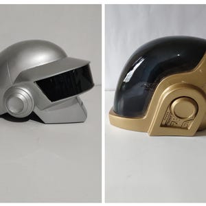 Może przedstawiać: Dwa futurystyczne hełmy, jeden srebrny i jeden złoty. Srebrny hełm ma czarną osłonę i okrągłe nauszniki. Złoty hełm ma czarną osłonę i szczeg&oacute;łowy panel boczny. Oba hełmy znajdują się na białej powierzchni.