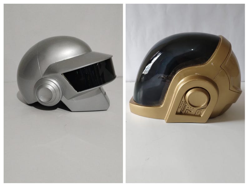 Może przedstawiać: Dwa futurystyczne hełmy, jeden srebrny i jeden złoty. Srebrny hełm ma czarną osłonę i okrągłe nauszniki. Złoty hełm ma czarną osłonę i szczeg&oacute;łowy panel boczny. Oba hełmy znajdują się na białej powierzchni.