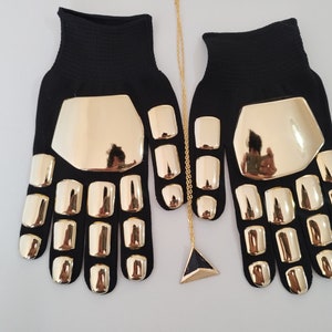 Puede incluir: Un par de guantes negros con detalles dorados. Los guantes tienen un diseño único con placas doradas que cubren los nudillos y los dedos. Un collar de cadena dorada con un colgante triangular cuelga debajo de los guantes.