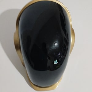 Może przedstawiać: Czarny hełm ze złotym obramowaniem. Hełm ma gładką, błyszczącą powierzchnię i zaokrąglony kształt. Złote obramowanie obrysowuje g&oacute;rę i boki hełmu, tworząc kontrastowy wz&oacute;r.