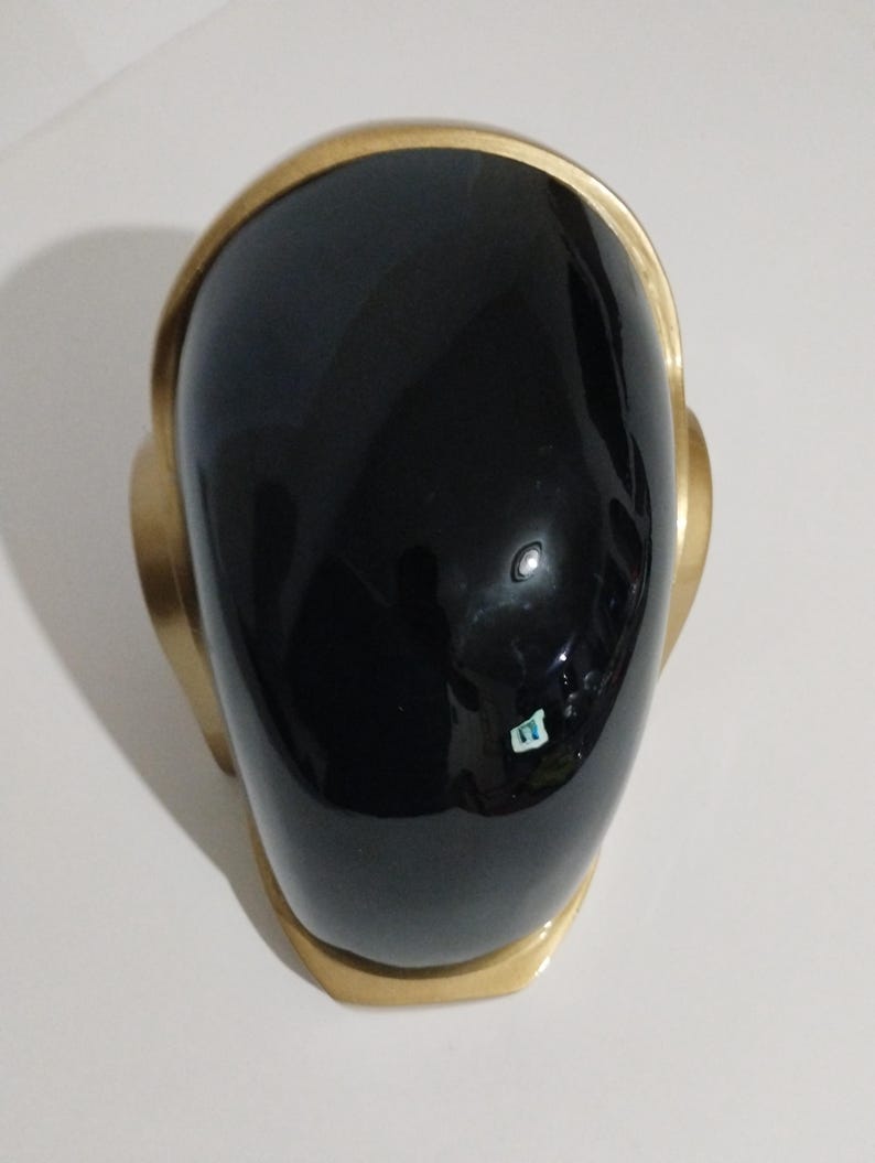 Może przedstawiać: Czarny hełm ze złotym obramowaniem. Hełm ma gładką, błyszczącą powierzchnię i zaokrąglony kształt. Złote obramowanie obrysowuje g&oacute;rę i boki hełmu, tworząc kontrastowy wz&oacute;r.