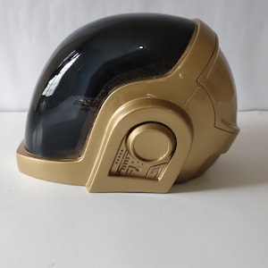 Może przedstawiać: Złoty kask z czarnym wizjerem. Kask ma gładkie, błyszczące wykończenie i okrągły detal z boku z szeregiem małych, prostokątnych i okrągłych wzor&oacute;w. Kask jest prawdopodobnie kostiumem lub przedmiotem kolekcjonerskim.