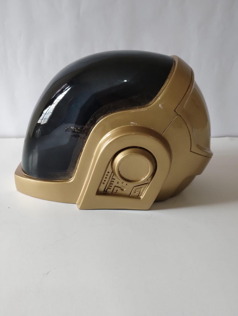 Może przedstawiać: Złoty kask z czarnym wizjerem. Kask ma gładkie, błyszczące wykończenie i okrągły detal z boku z szeregiem małych, prostokątnych i okrągłych wzor&oacute;w. Kask jest prawdopodobnie kostiumem lub przedmiotem kolekcjonerskim.