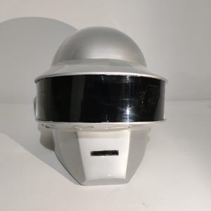 Może przedstawiać: Srebrno-czarny hełm z zaokrągloną g&oacute;rą i prostokątną twarzą. Hełm ma czarną osłonę i mały prostokątny otw&oacute;r na twarzy. Hełm jest prawdopodobnie kostiumem lub rekwizytem.