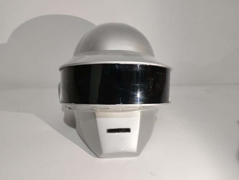 Może przedstawiać: Srebrno-czarny hełm z zaokrągloną g&oacute;rą i prostokątną twarzą. Hełm ma czarną osłonę i mały prostokątny otw&oacute;r na twarzy. Hełm jest prawdopodobnie kostiumem lub rekwizytem.