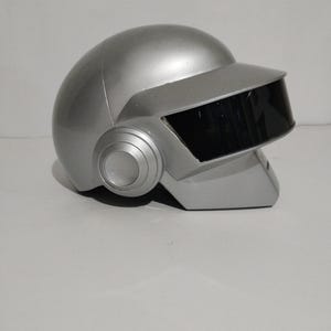 Może przedstawiać: Srebrny kask z czarnym wizjerem i okrągłymi nausznikami. Kask ma gładkie, metaliczne wykończenie i futurystyczny design. Wizjer jest prostokątny i zakrywa obszar twarzy. Projekt przypomina artyst&oacute;w muzyki elektronicznej.
