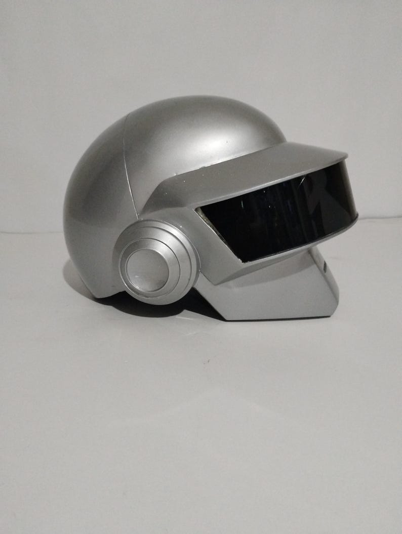 Może przedstawiać: Srebrny kask z czarnym wizjerem i okrągłymi nausznikami. Kask ma gładkie, metaliczne wykończenie i futurystyczny design. Wizjer jest prostokątny i zakrywa obszar twarzy. Projekt przypomina artyst&oacute;w muzyki elektronicznej.