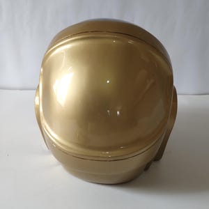 Może przedstawiać: Złoty kask o gładkiej, odblaskowej powierzchni. Kask ma zaokrąglony kształt z obszarem wizjera i elementami bocznymi, sugerującymi futurystyczny lub kosmiczny design. Og&oacute;lna estetyka jest elegancka i nowoczesna.