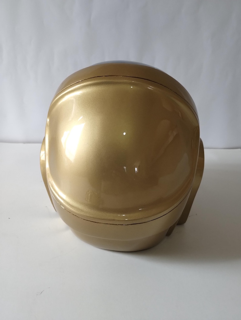 Może przedstawiać: Złoty kask o gładkiej, odblaskowej powierzchni. Kask ma zaokrąglony kształt z obszarem wizjera i elementami bocznymi, sugerującymi futurystyczny lub kosmiczny design. Og&oacute;lna estetyka jest elegancka i nowoczesna.