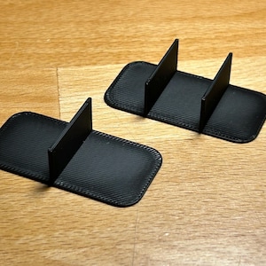 以下が含まれることがあります： 黒色の長方形の電話スタンド2つ。表面はテクスチャ加工され、三角形のサポートが付いています。木製の表面に配置され、ミニマリストなデザインを示しています。各スタンドには1つのサポートがあります。