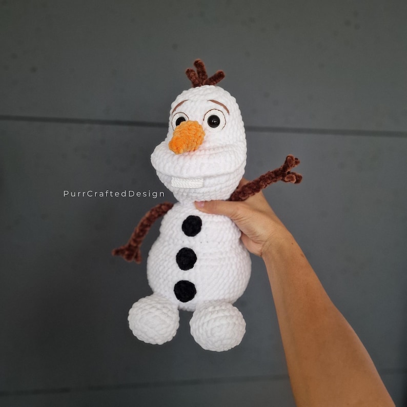 May include: A white crochet snowman with a carrot nose, black button accents, and brown twig arms and hair. The snowman has a friendly expression and is a handmade item.