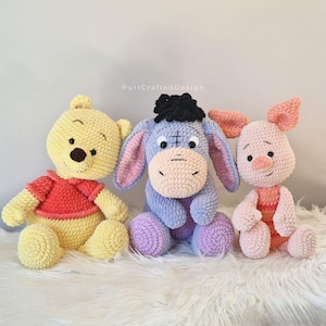 Pode incluir: Tr&ecirc;s brinquedos de pel&uacute;cia de croch&ecirc; feitos &agrave; m&atilde;o: um urso amarelo vestindo uma camisa vermelha, um burro azul com cabelo preto e um porco rosa. Os brinquedos est&atilde;o sentados em uma superf&iacute;cie branca. O urso tem um nariz marrom e o porco tem um focinho rosa.