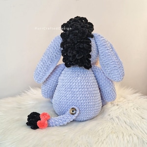 Pode incluir: Um brinquedo de pel&uacute;cia de croch&ecirc; azul claro com uma crina preta e uma cauda com detalhes em preto e coral. O brinquedo de pel&uacute;cia tem um bot&atilde;o na parte de tr&aacute;s e est&aacute; sentado em uma superf&iacute;cie branca e fofa. O texto "PurrCraftedDesign" &eacute; vis&iacute;vel.