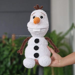 May include: A handmade white crochet snowman doll with a carrot nose, black button accents, and brown twig arms and hair. The doll has a smiling face and is being held by a person. The text "PurrCraftedDesign" is visible.