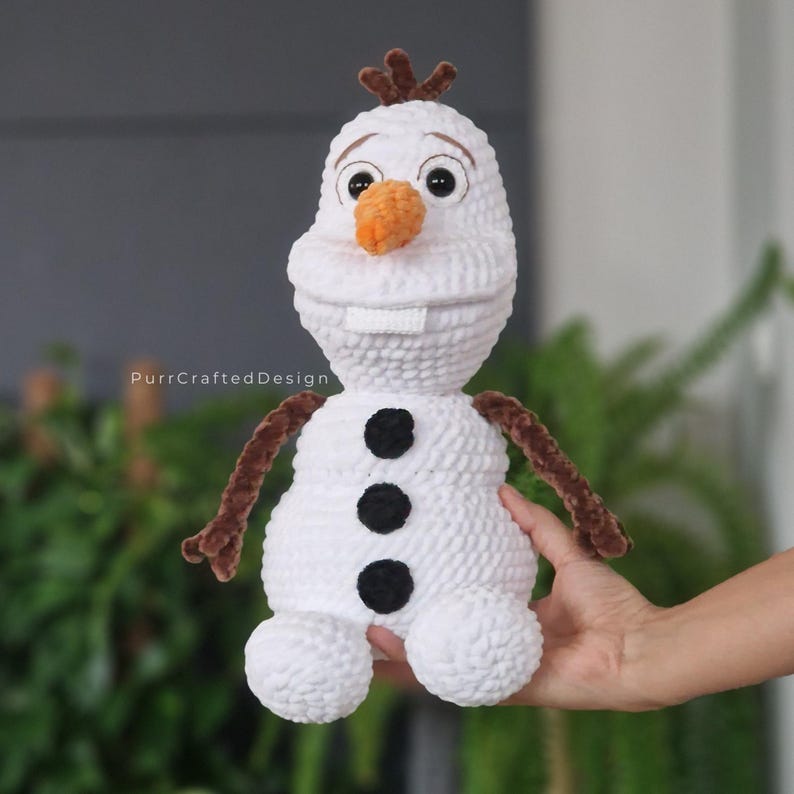 May include: A handmade white crochet snowman doll with a carrot nose, black button accents, and brown twig arms and hair. The doll has a smiling face and is being held by a person. The text "PurrCraftedDesign" is visible.