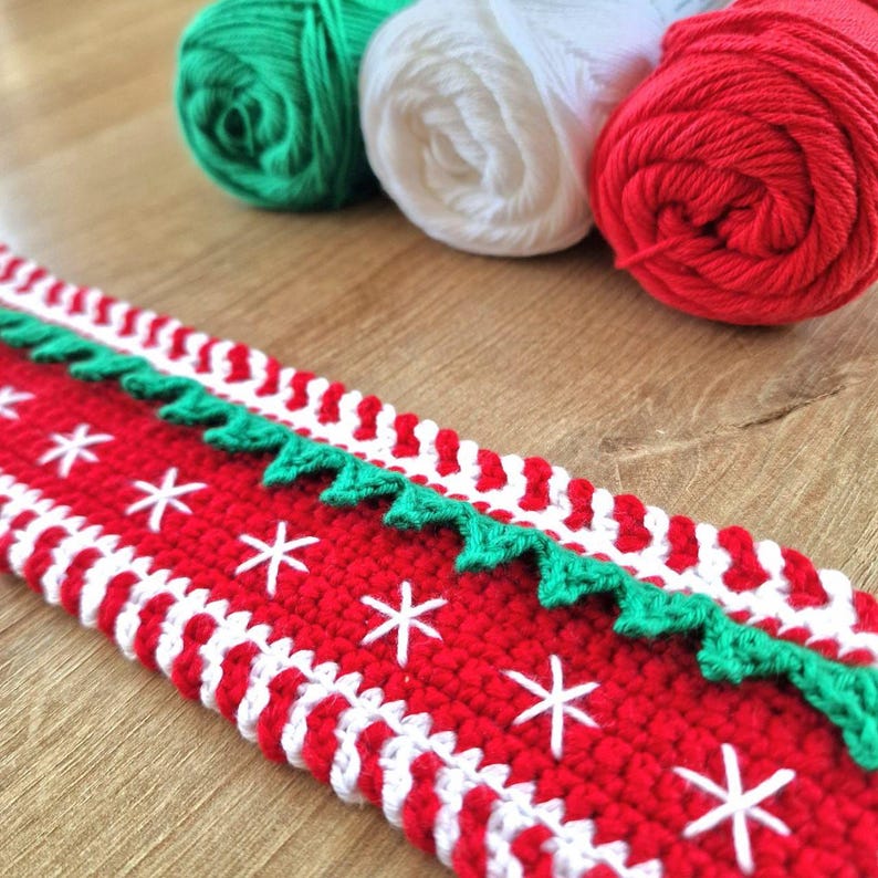 May include: A red, white, and green crocheted Christmas garland with a snowflake pattern. The garland is made with yarn and has a scalloped edge.