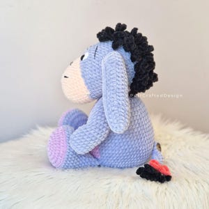 Pode incluir: Um brinquedo de pel&uacute;cia feito &agrave; m&atilde;o de Eeyore, o burro de Winnie the Pooh. A pel&uacute;cia &eacute; principalmente azul claro com um focinho e p&eacute;s rosa. Possui uma crina e cauda pretas e est&aacute; sentado em uma superf&iacute;cie branca e fofa. O brinquedo &eacute; de croch&ecirc;.