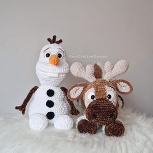 May include: Two crocheted plush toys: a white snowman with a carrot nose and black buttons, and a brown reindeer with large antlers. The snowman has brown arms and the reindeer has white accents. The toys are handmade.