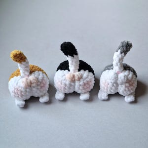 Può includere: Tre peluche fatti a mano a forma di sedere di gatto in fila. I peluche sono bianchi con accenti rosa e ognuno ha una coda di colore diverso: arancione e bianco, nero e bianco e grigio e bianco. I peluche sono fatti di filato morbido e strutturato.