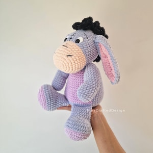 Pode incluir: Um brinquedo de pel&uacute;cia Eeyore feito &agrave; m&atilde;o em croch&ecirc;. O burro tem um corpo roxo claro, uma barriga rosa e um focinho bege. As orelhas s&atilde;o roxas claras com o interior rosa, e a crina &eacute; preta.