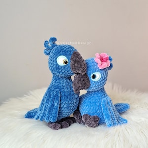 May include: Two plush blue bird toys with gray beaks and feet. One bird has a pink flower on its head. The birds are sitting on a white, fluffy surface. The birds are made of a soft, textured material.