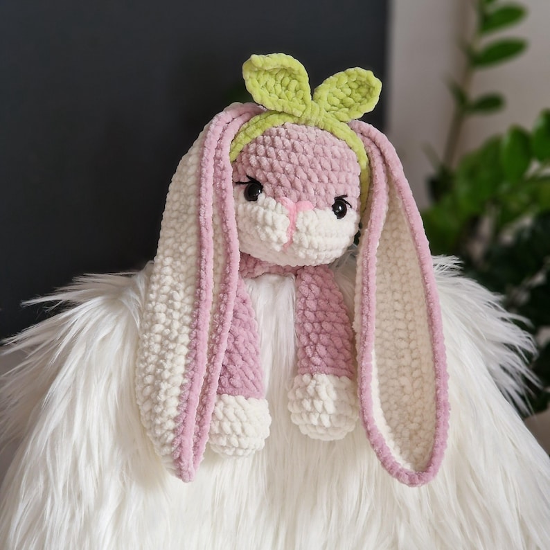 May include: A pink and white crocheted bunny with long ears and a green bow. The bunny is sitting on a white fluffy surface.