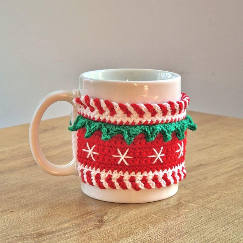 May include: White ceramic mug with a red and white crocheted mug cozy featuring a green trim and white snowflakes.