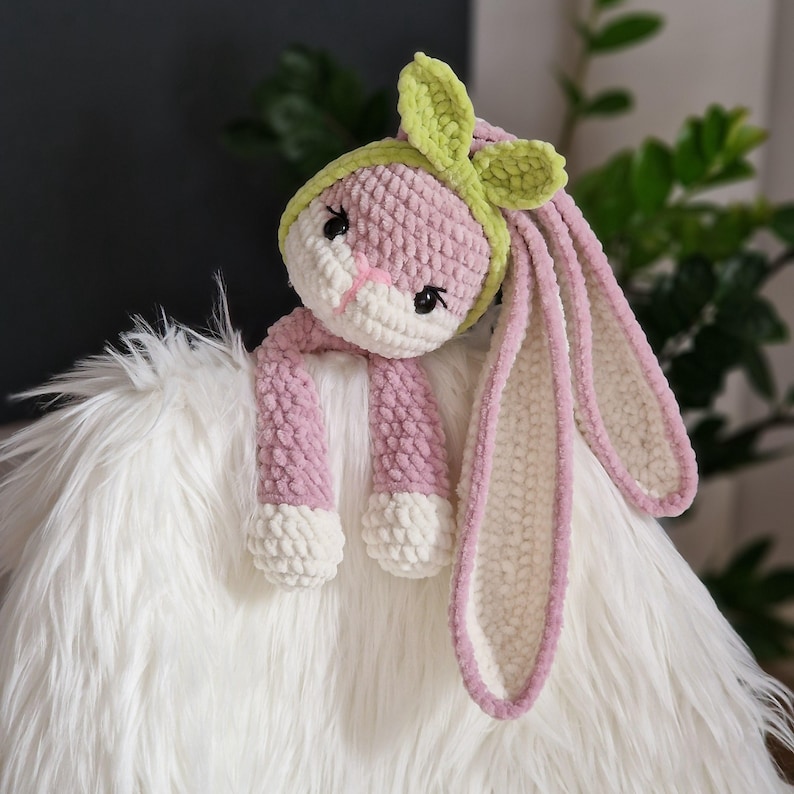 May include: A pink and white crocheted bunny with long ears and a green bow. The bunny is sitting on a white fluffy surface.