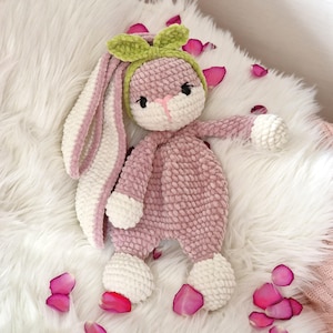 May include: A pink crocheted bunny with white accents and a green bow. The bunny is sitting on a white fluffy surface with pink flower petals.