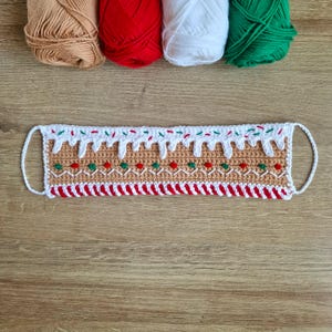 May include: A crocheted gingerbread house face mask with white icing, red and green candy sprinkles, and a red and white candy cane border.