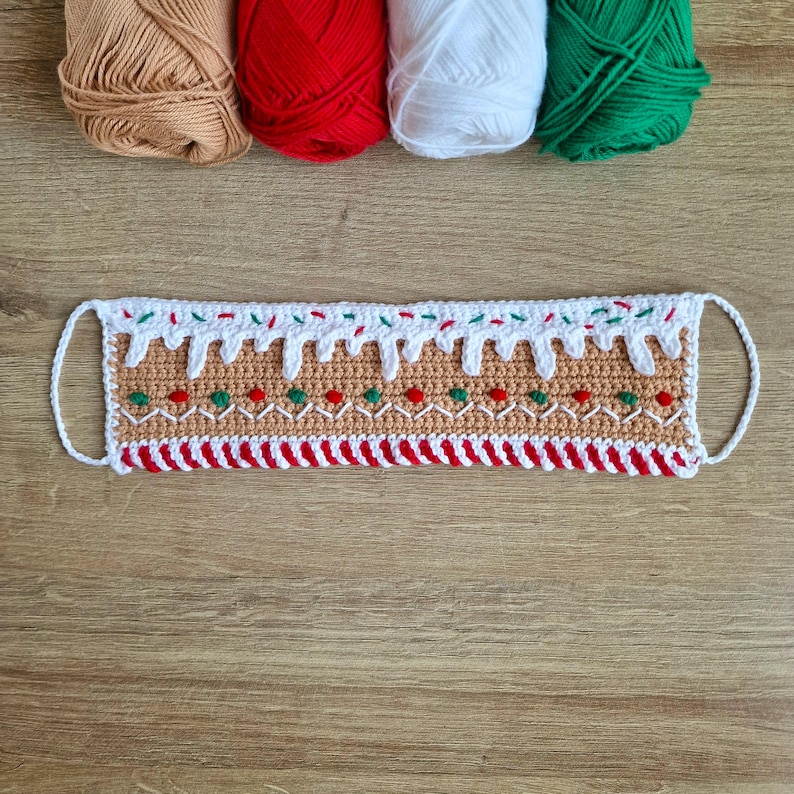 May include: A crocheted gingerbread house face mask with white icing, red and green candy sprinkles, and a red and white candy cane border.